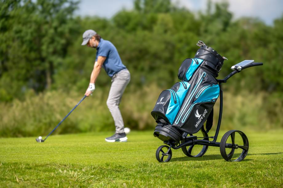 Ein Golfspieler beim Golfen, im Vordergrund steht ein Golftrolley mit Golf-Bag